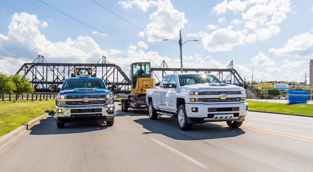 A white and a blue Chevy Silverado 2500 HD both towing heavy machinery
